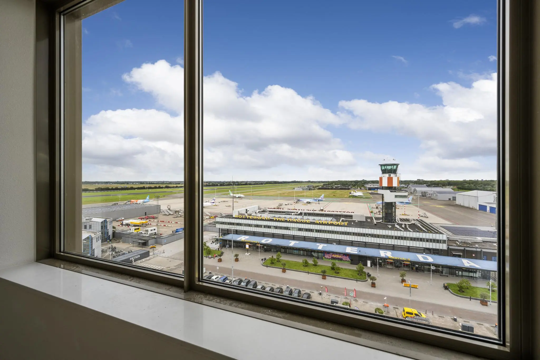 Uitzicht door een raam op Rotterdam The Hague Airport met terminal, verkeerstoren en geparkeerde vliegtuigen.